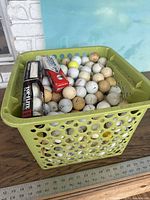 Top-angle photo of green plastic laundry basket full of mostly used and some new golf balls, including tubes of branded golf balls Top-Flite and Dura-Wear.