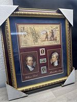 Framed poster art with gold-tone frame, burgundy mat, vintage map, portraits of William Clark and Meriwether Lewis, and replica postage stamps.