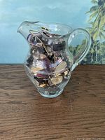 Glass pitcher filled with assorted jewelry items including watches, bracelets, cufflinks, and necklaces.