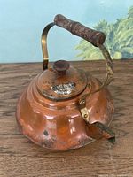 Close-up showing the full copper teapot with wooden handle and spout on a wooden surface, clearly showing wear and patina.
