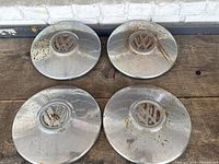 Four Volkswagen hubcaps placed on wooden surface showing varying levels of surface rust and wear around edges and logos.