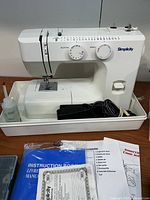 Front view of white Simplicity sewing machine on tray with power cord, foot pedal, small oil bottle, instruction manual booklet, and accessory paperwork.