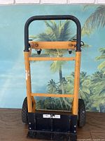 Front view of the small multipurpose dolly showing the orange-yellow frame, black handle, and wheels against a tropical background.