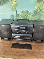 Sony CFD-565 boombox showing full front with dual speakers and detached cassette cover on wood surface with tropical backdrop.