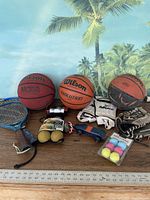 Full lot photo showing all sports items including basketballs, rackets, gloves, balls, and accessories arranged on a wooden surface with a tropical background