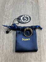 Ring resting on navy blue satin Stauer pouch with gold logo and attached tag, showing the band and stones.