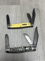 Photo showing two vintage pocket knives laid open on floor, one with yellow handle and three blades, one with textured black and white handle and two blades.