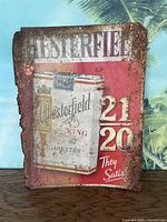 Front view of vintage metal advertisement sign for Chesterfield King cigarettes showing cigarette pack image, text, and heavily rusted edges.
