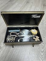 Open brass trinket box showing assorted vintage sewing supplies inside including button hooks, thread spools, scissors, and small figurines.