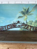 Full view of the metal hanging sign with rustic finish placed against a backdrop showing tropical imagery.