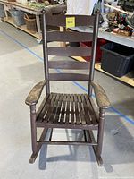 Front view of a dark brown wooden rocking chair with a slatted seat and backrest showing rustic wear on armrests.