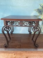Full front view of the wooden top metal end table showing the rectangular wooden surface and detailed curved metal legs with ornate scrollwork along the apron.