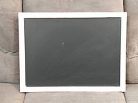 Photo of a rectangular chalk board with a white wood frame, resting on a beige fabric surface. Frame shows minor wear.