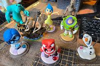 Photo showing eight Disney Infinity character figures on a surface including Buzz Lightyear, Olaf, Sulley, Joy, Sadness, Anger, Groot, and a pirate ship figure