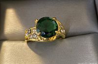 Close-up photo of gold tone ring with large green stone and small clear stones on band.