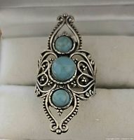 Close-up photo of silver toned ring facing front, showing detailed scrollwork design and three round blue stones of varying sizes.