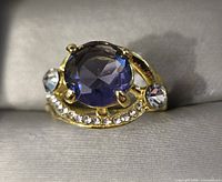 Close-up photo of gold toned ring with large oval blue stone and clear accent stones on a curved band