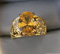 Front view of gold toned fashion ring with large oval yellow gemstone and clear stones on openwork band.