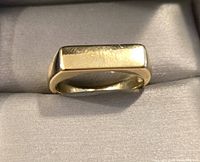 Gold toned solid ring placed on light gray cushioned fabric inside a box, showcasing the flat rectangular top and polished band.