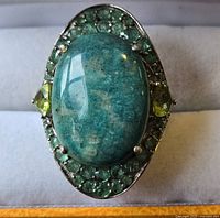 Front view of large silver-toned ring with oval blue-green stone centerpiece surrounded by multiple smaller green stones and two triangular yellow-green stones on the sides.