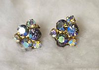 Close-up photo of a pair of small clip-on earrings with multicolored rhinestones on gold-tone clip-on backs, displayed on a soft white surface.