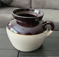 Photo of Robinson Ramsbottom pottery bean pot, showing two-tone dark brown and light ceramic glaze, rounded body with handle, pot on black table in front of couch