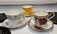 Three teacup and saucer sets displayed together with floral and gold designs.