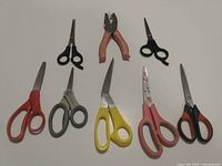 Seven items shown on white background including six pairs of scissors with various handle colors and one metal hole punch tool in center with pink grips.