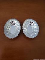 Two small sterling silver dishes with oval shapes and scalloped edges, placed on a wooden surface.