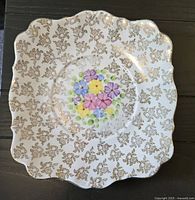 Top view of square bone china cake plate showing colorful floral design in center with gold leaf pattern on white base and scalloped, gold-trimmed edges.