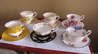 Seven assorted fine china teacups and saucers arranged showing cup sides and saucer patterns, including a mismatched Royal Albert cup and Shafford saucer.