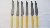Six dinner cutting knives with yellow handles and stainless blades laid side by side on a white surface.