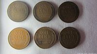 Six Lincoln Wheat pennies arranged in two rows on a white surface showing the reverse side with 'ONE CENT' and wheat stalks.