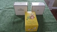 Photo showing two white Chanel No. 5 perfume boxes and one yellow Laura Secord Easter egg cardboard box on a green surface.