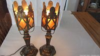 Pair of vintage brass lamps lit with amber-colored shades glowing, showing ornate brass frames with floral and female figure decorations.