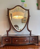 Frontal view of shield-shaped beveled dressing mirror on wooden base with three drawers.