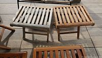 Two teak wooden adjustable foot stools on wooden outdoor flooring, showing natural weathering and patina of wood.