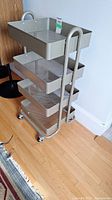 Overall view of the gray metal cart with four mesh shelves and wheels on hardwood floor.