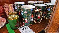 Photo showing six Christmas-themed mugs with gold handles and two green candle holders on a wooden shelf.