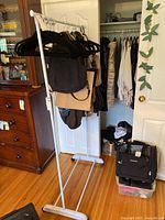 White metal rolling clothing rack with black hangers holding bags, positioned on wooden floor indoors.