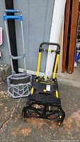 Two dollies side by side outside near a shed door, Stanley dolly in black and yellow and a gray metal/plastic dolly with blue handles