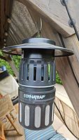 Front view of the Dynatrap mosquito repeller hanging outdoors against wood and greenery background. Shows ventilation slits, mesh trap area, and top hat-shaped cover. Device is black with white logo text and branding on the central band.