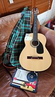 Photo showing full guitar body with soft plaid guitar case and guitar lesson book with CD on a brown leather couch.