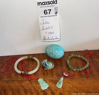 Full lot shot showing two jade bracelets, two carved jade pendant charms on red cords, and two decorative eggs on wooden surface beneath auction tag.