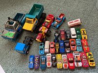 Photo shows a large assortment of toy and model cars and trucks arranged on carpet, including two large dump trucks, several small diecast cars, and vintage toy cars.