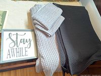 Two blankets, light gray knit pattern and dark gray/black quilted, with a framed "Stay Awhile" sign on wood.