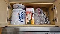 View of cabinet shelf showing stacks of Royal Chinet paper plates and cups, Foodsaver replacement rolls box, and metal wire utensil holder.