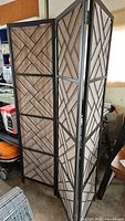 Wide angle photo showing the room divider partially extended, displaying all three panels with geometric patterned wood-textured panels and black metal frames in a storage or garage setting.
