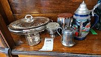 Photo showing six items: metal and glass casserole dish with embossed lid, ceramic tankard with lid, small metal pitcher, glass flower frog in metal holder, two metal candle holders, and a tin container.