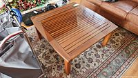 Full view of square teak coffee table with slatted top and removable glass cover on patterned rug